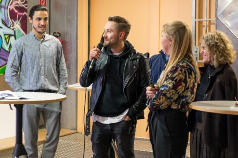 Mehrere Personen im Bild.. Ein Mann spricht ins Mikrofon.