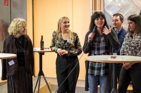 Mehrere Personen im Bild.. Eine Frau spricht ins Mikrofon.