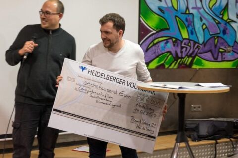 Eine Person am Mikrofon, eine hält den Scheck mit dem Wert von 6000 Euro